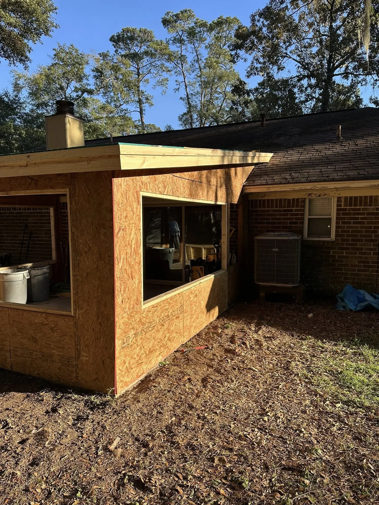 Backyard construction of a small extension or room attached to a brick house, with unfinished wood siding and window, on a dirt ground with construction tools and materials.