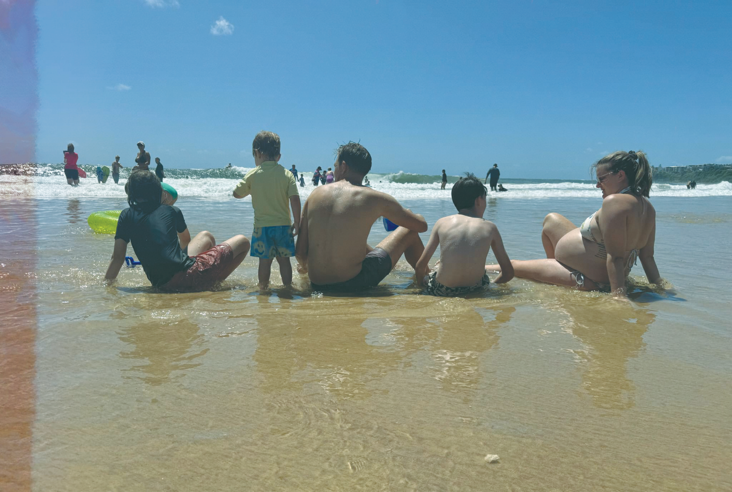 A group of people, including children and adults, sitting and relaxing in shallow water at the beach under a bright blue sky with some clouds, with more people swimming and playing in the ocean in the background.