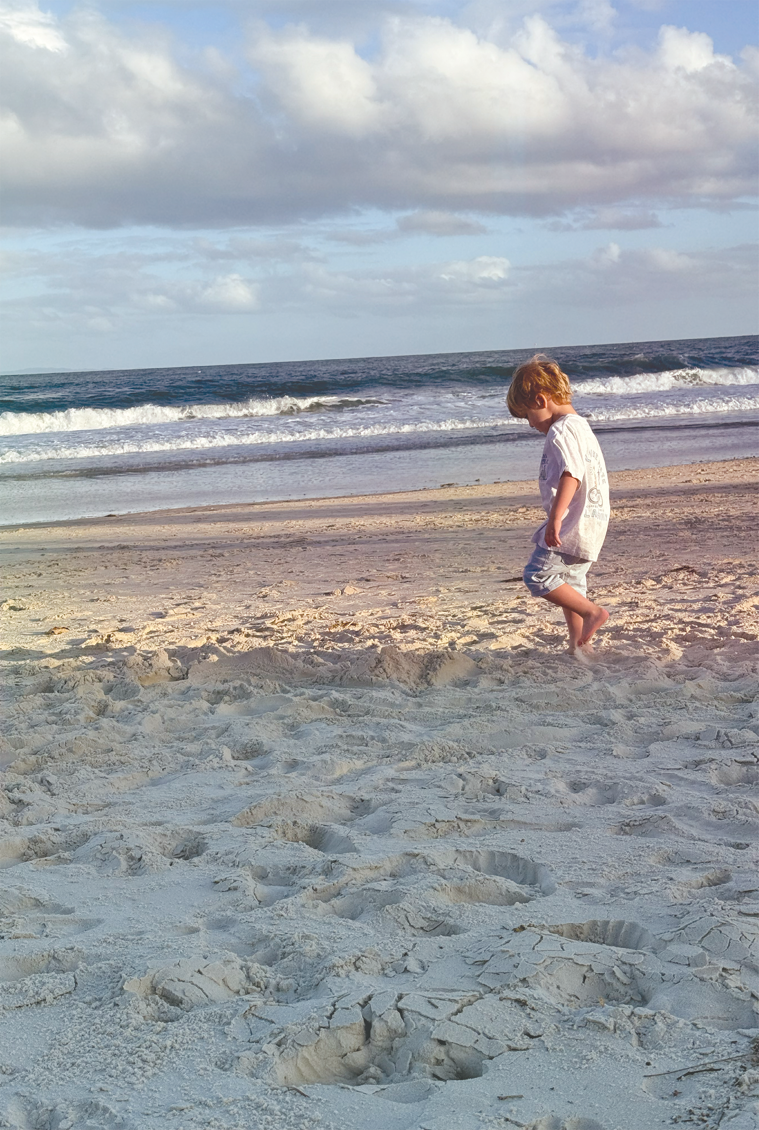 A young boy with blonde hair walking barefoot on the sandy beach near the water. The sky is partly cloudy, and gentle waves are approaching the shore.