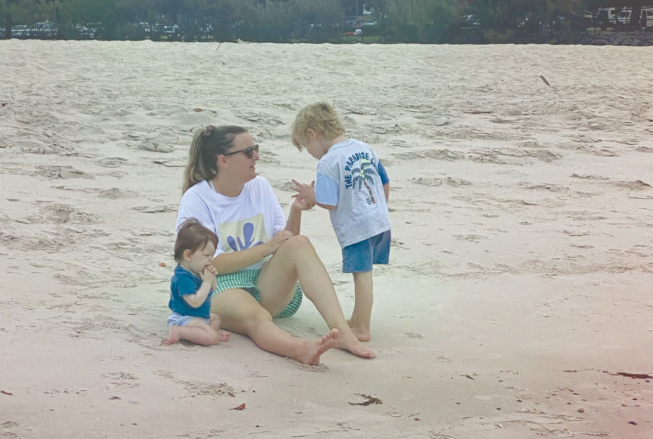 A woman with two children sitting on the beach, one child is sitting on her lap and the other is standing and talking to her, in a sandy area with water in the background.