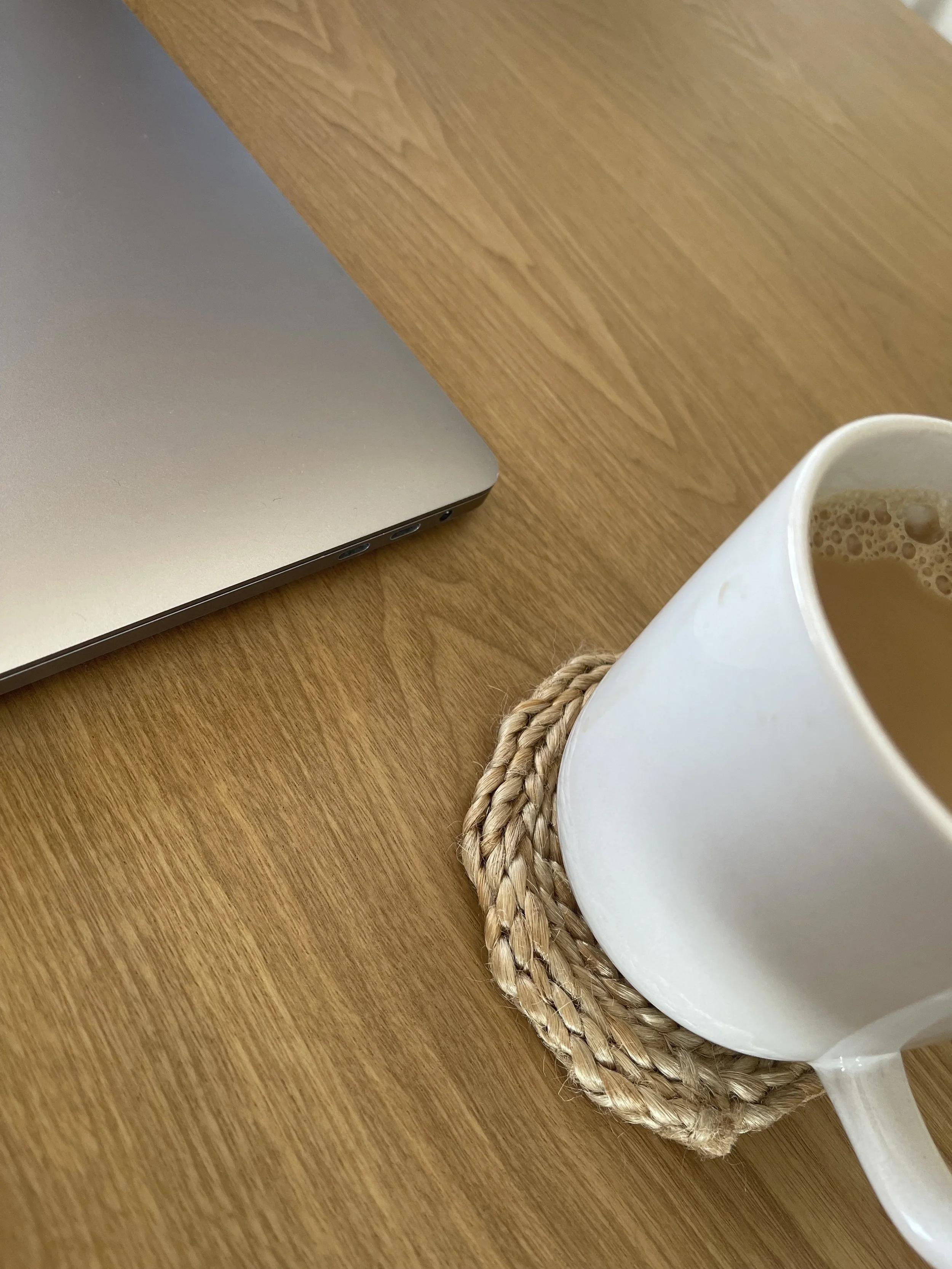A laptop on a wooden desk, next to a mug of coffee on a woven coaster.
