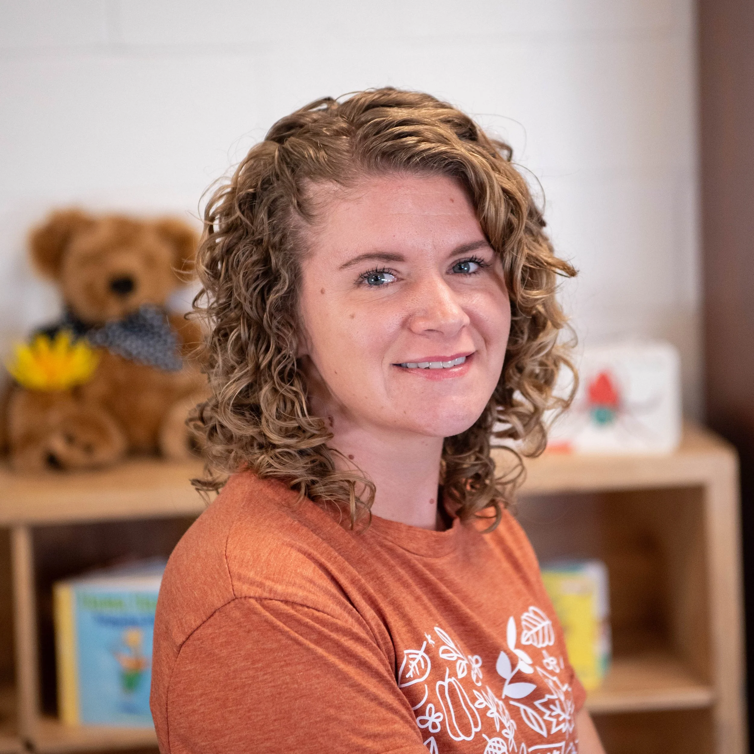 A woman with curly hair wearing an orange shirt, smiling, with a brown teddy bear and books in the background.
