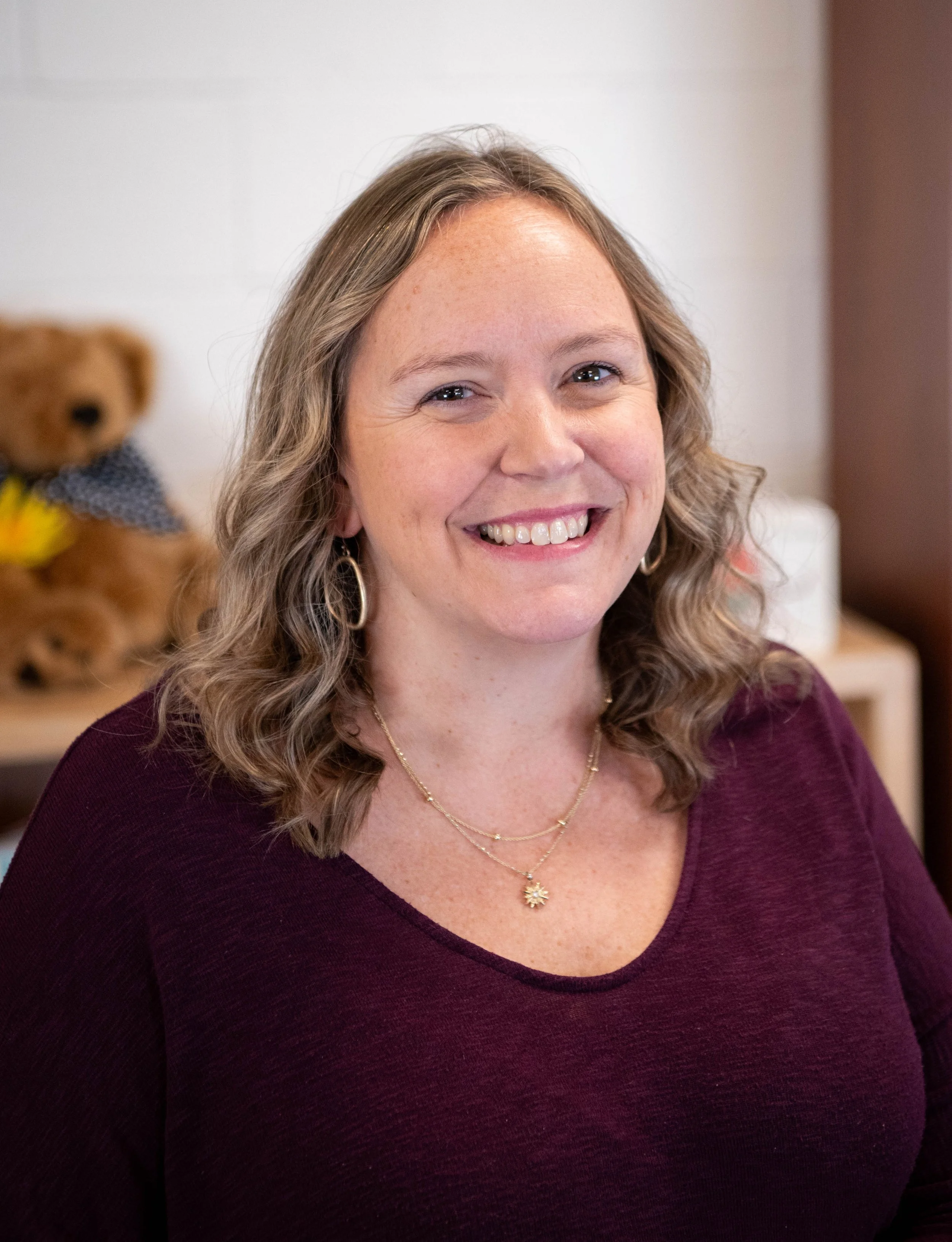 A woman with shoulder-length wavy hair wearing a maroon top, gold hoop earrings, and layered gold necklaces, smiling at the camera with a teddy bear in the background.