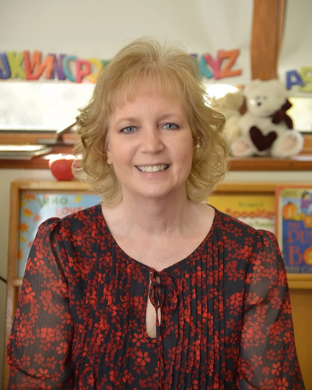 A woman with shoulder-length blonde hair and blue eyes smiling, sitting in a classroom or children's reading area with colorful books, stuffed animals, and alphabet letters in the background.
