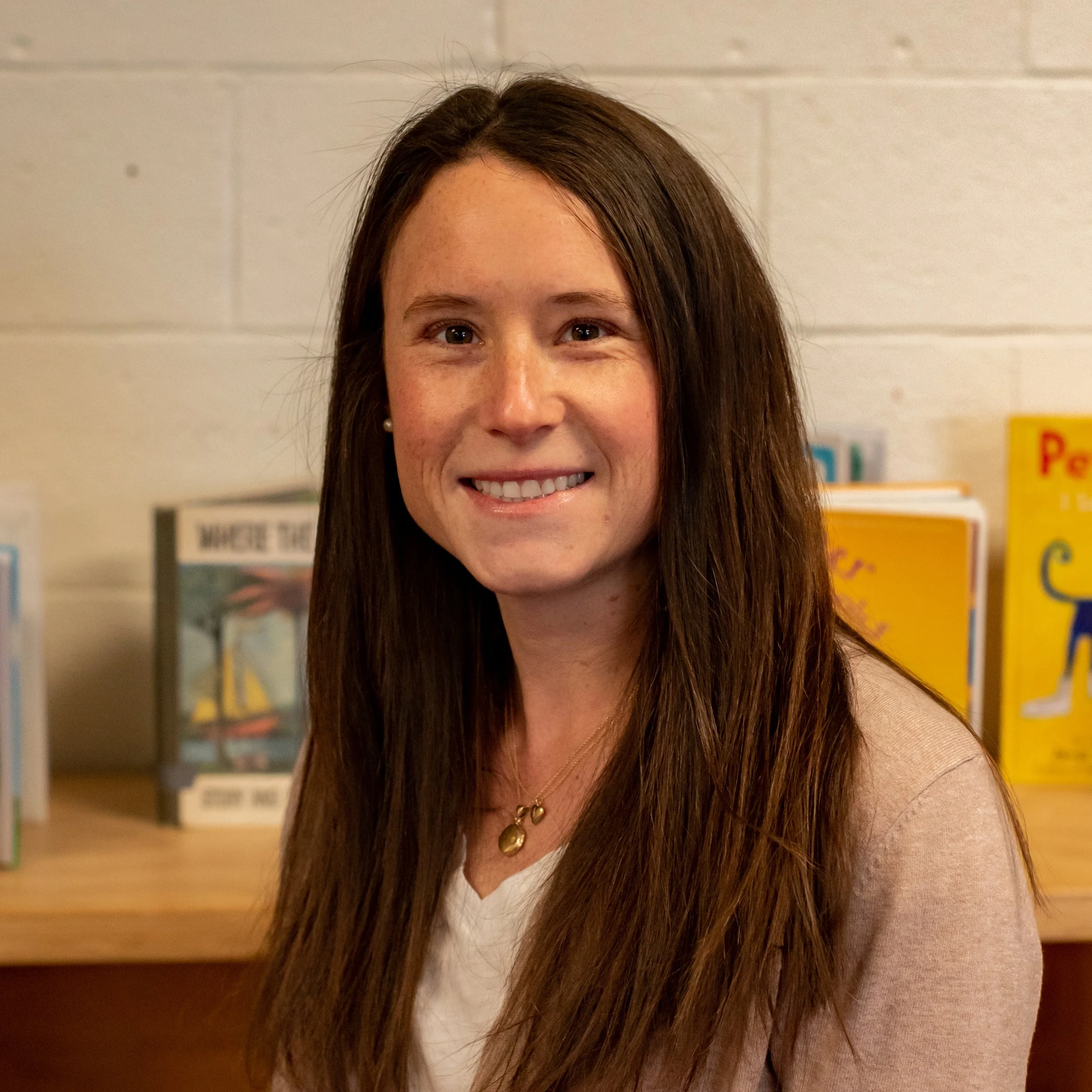 A smiling woman with long brown hair wearing a white top and gold jewelry, standing in front of a bookshelf filled with children's books.