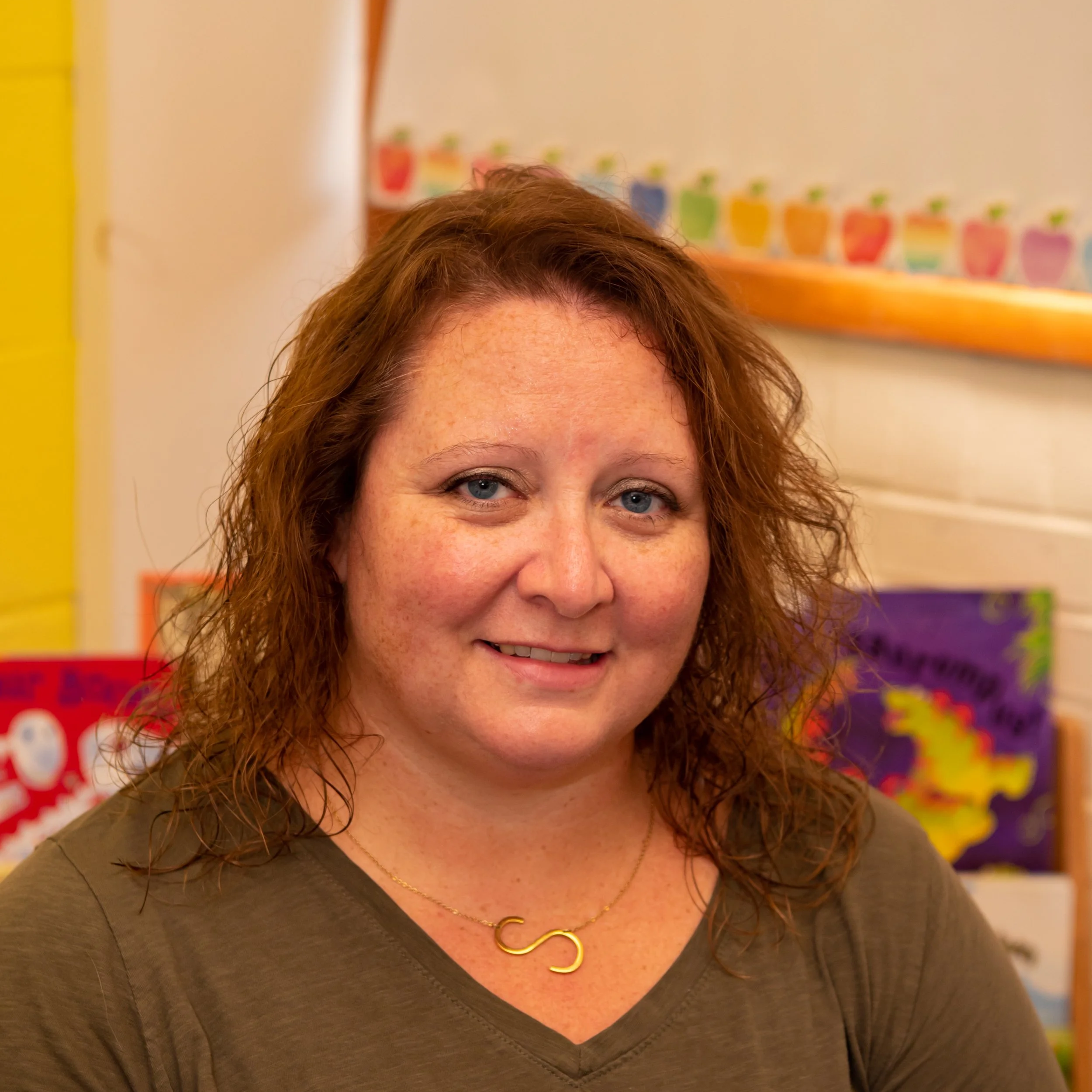 A woman with curly red hair smiling in a colorful classroom with children's artwork on the wall behind her.