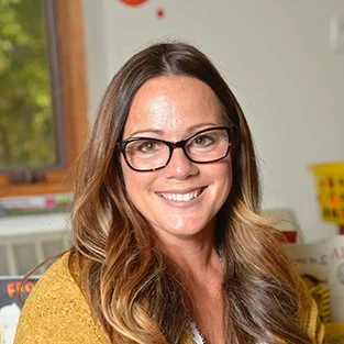 Smiling woman with glasses and wavy brown hair tagged in a room with a window and colorful objects in the background.