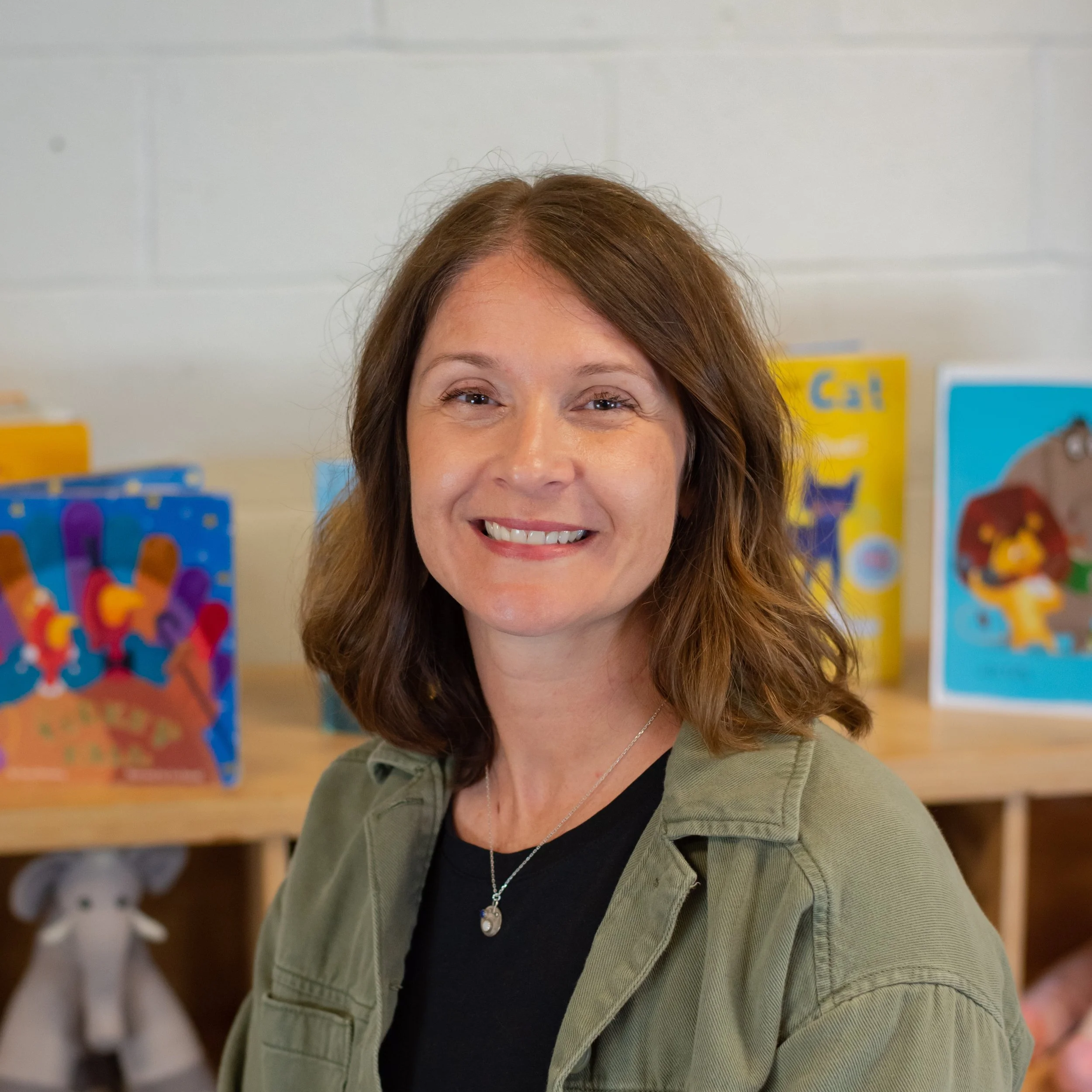 A woman with shoulder-length brown hair smiling in front of children's books on a shelf.
