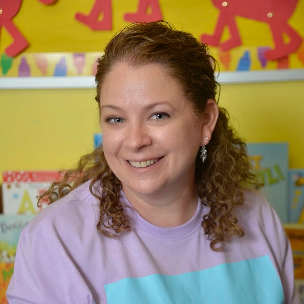 A woman with brown curly hair and earrings smiling at the camera in front of a colorful classroom display.