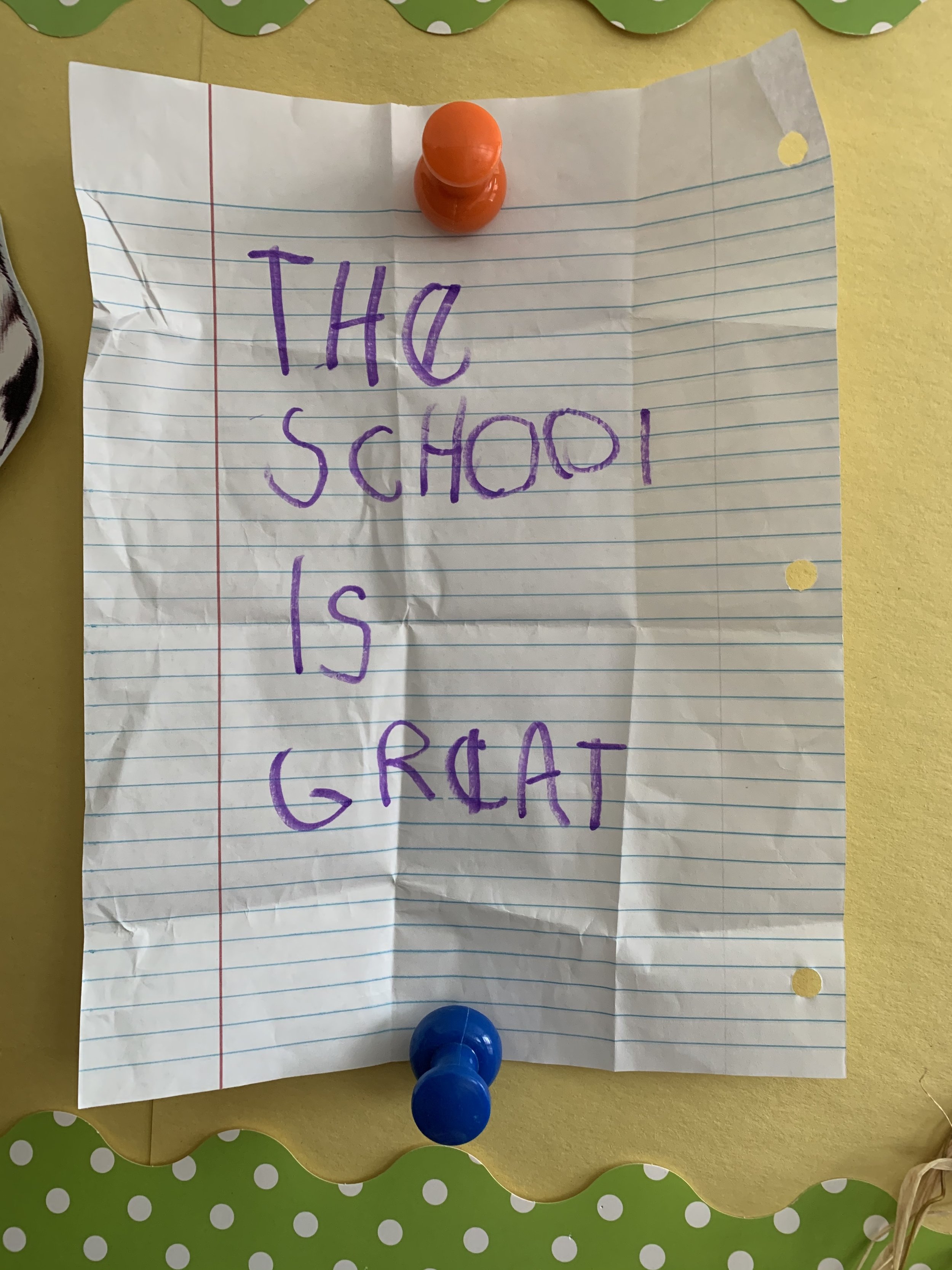 A crumpled sheet of lined notebook paper with the handwritten message 'The school is great' in purple marker, pinned to a yellow bulletin board with red and blue push pins.