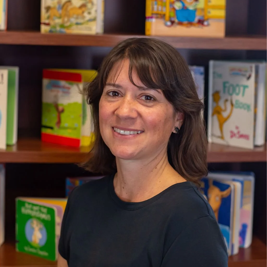 A woman with short brown hair and a black shirt smiling in front of a bookshelf filled with children's books.