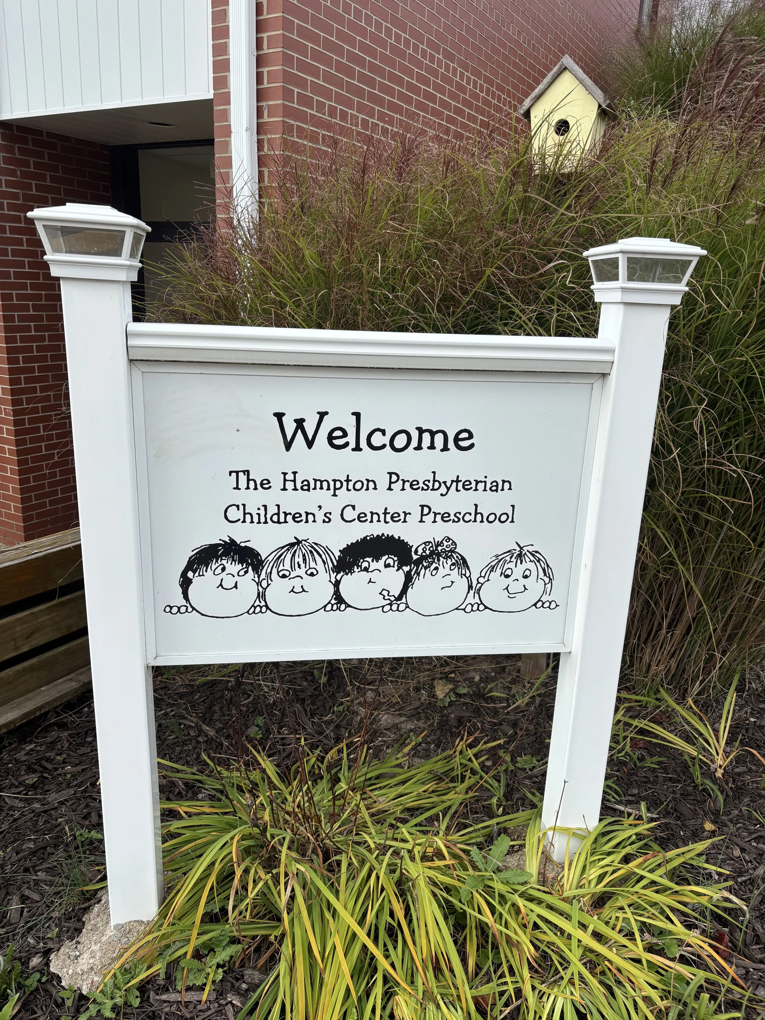 White welcoming sign for The Hampton Presbyterian Children's Center Preschool with cartoon children drawings, set in a bed of plants and tall grass, with a birdhouse on a brick building in the background.