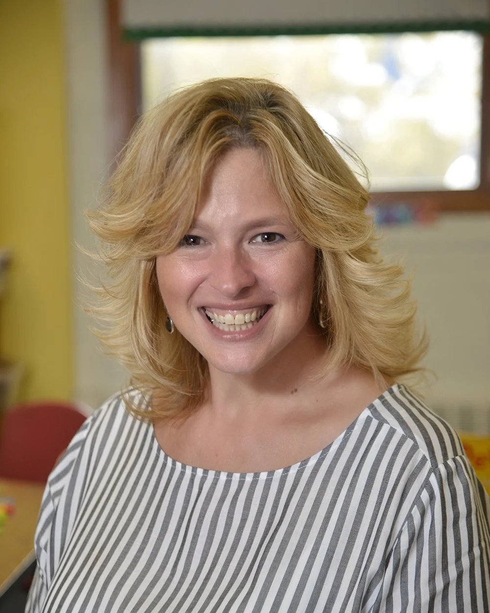 Portrait of a woman with blonde hair smiling, wearing a striped shirt, indoors with a blurred background.