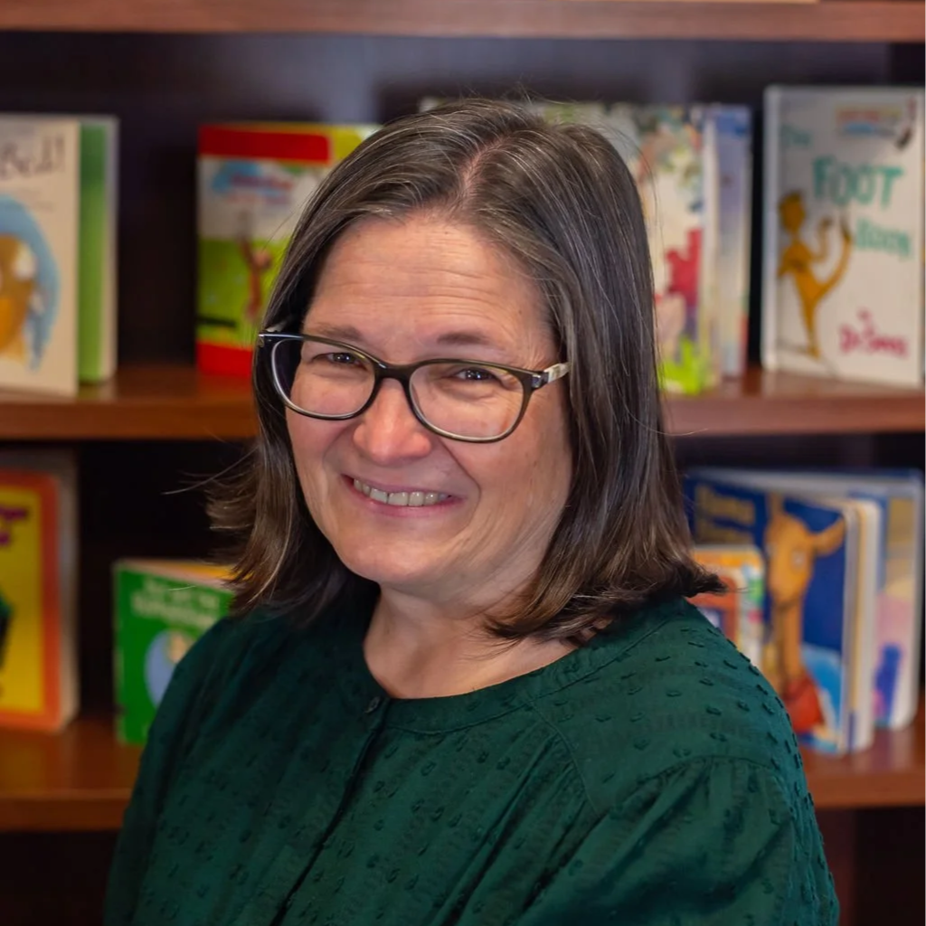 A woman with shoulder-length brown hair and glasses smiling in front of a bookshelf filled with children's books.