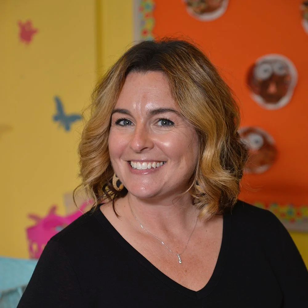 A woman with shoulder-length wavy hair smiling in front of a colorful background with cartoonish decorations and faces.