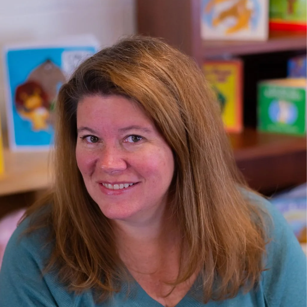 A woman with shoulder-length red hair smiling at the camera, in a room with books and colorful children's illustrations in the background.