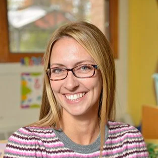 A woman with blonde hair and glasses smiling indoors, with a colorful background that appears to be a classroom or children's area.