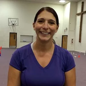 A woman with dark hair pulled back, smiling, wearing a purple athletic shirt, standing inside a gym or sports facility with a cross on the wall.