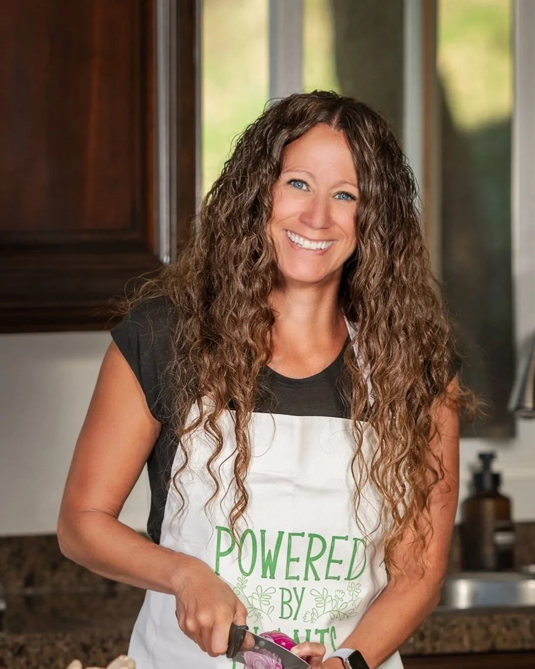 A smiling woman with long curly hair wearing a white apron that says 'Powered by Plant Based' over a black shirt, chopping purple onion in a kitchen with wooden cabinets and a window.