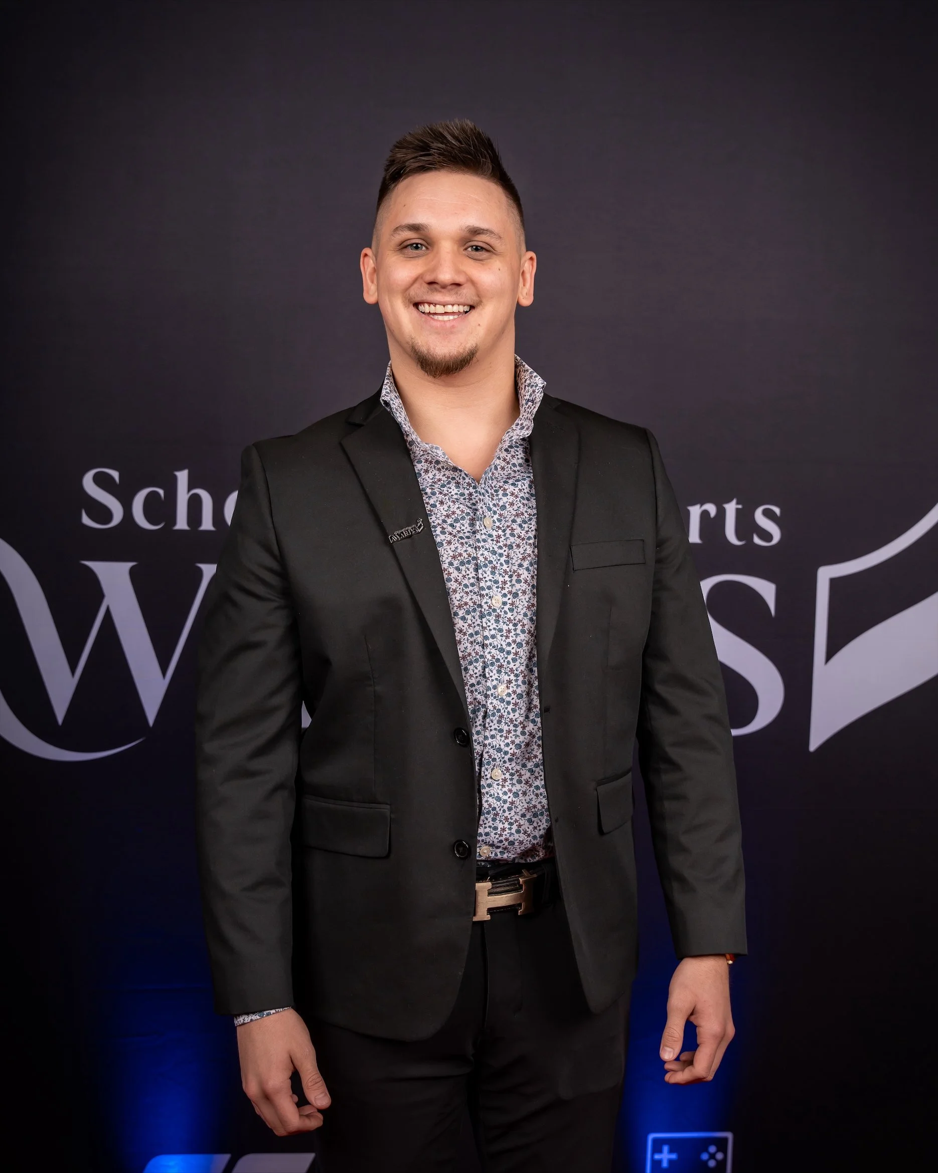 A young man in a black business suit with a patterned shirt smiling in front of a dark background.