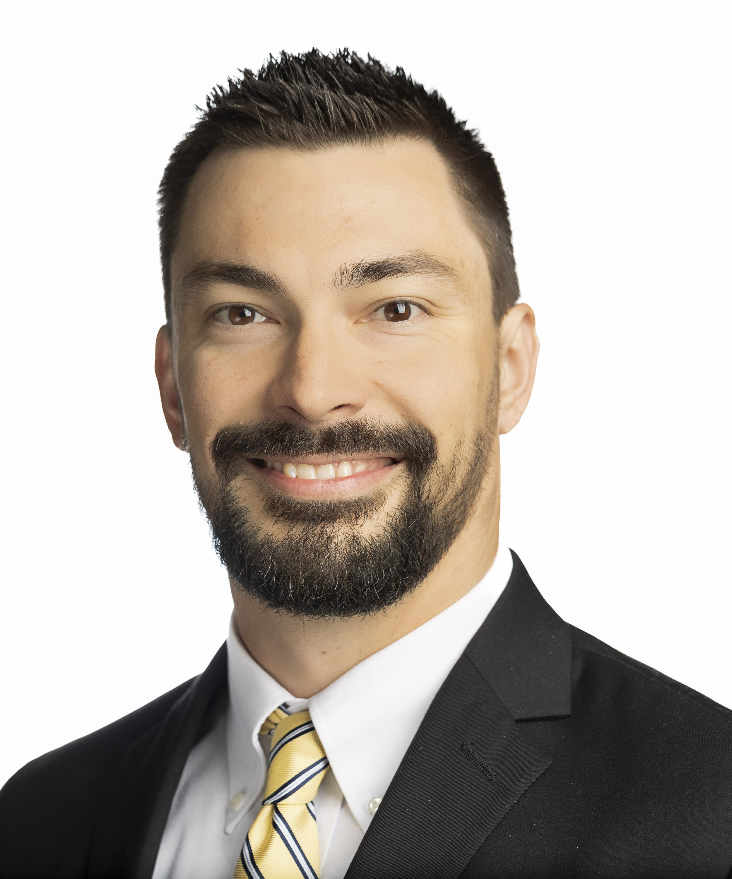 Close-up of a smiling man with a beard in a formal suit and striped yellow tie against a white background.