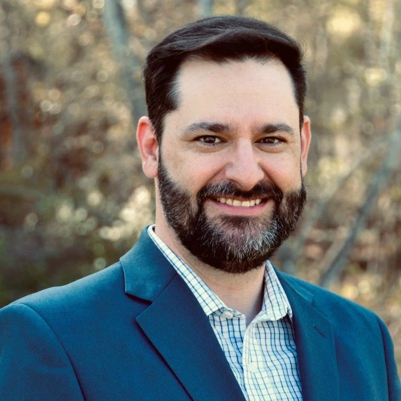 A smiling man with dark hair and a beard, wearing a blue suit jacket and a checkered shirt, outdoor background with blurred trees.
