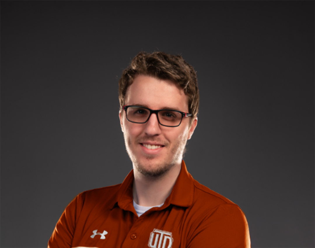 Headshot of a young man with glasses and a slight smile, wearing a burnt orange Under Armour polo shirt with a university logo, against a dark gray background.