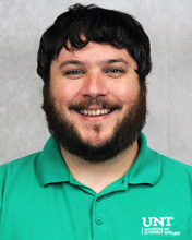 Portrait of a man with dark hair and a beard, smiling, wearing a teal polo shirt, against a plain gray background.