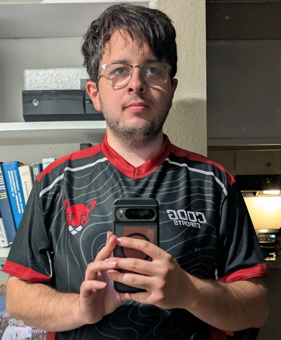 A man with glasses and dark hair taking a selfie in a mirror, wearing a black and red sports jersey with a lion logo and the text 'COG' and 'ESPORTS' on it.