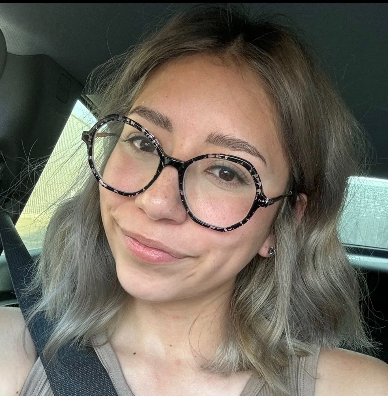 A young woman with glasses in a car, smiling at the camera with wavy hair and natural makeup.