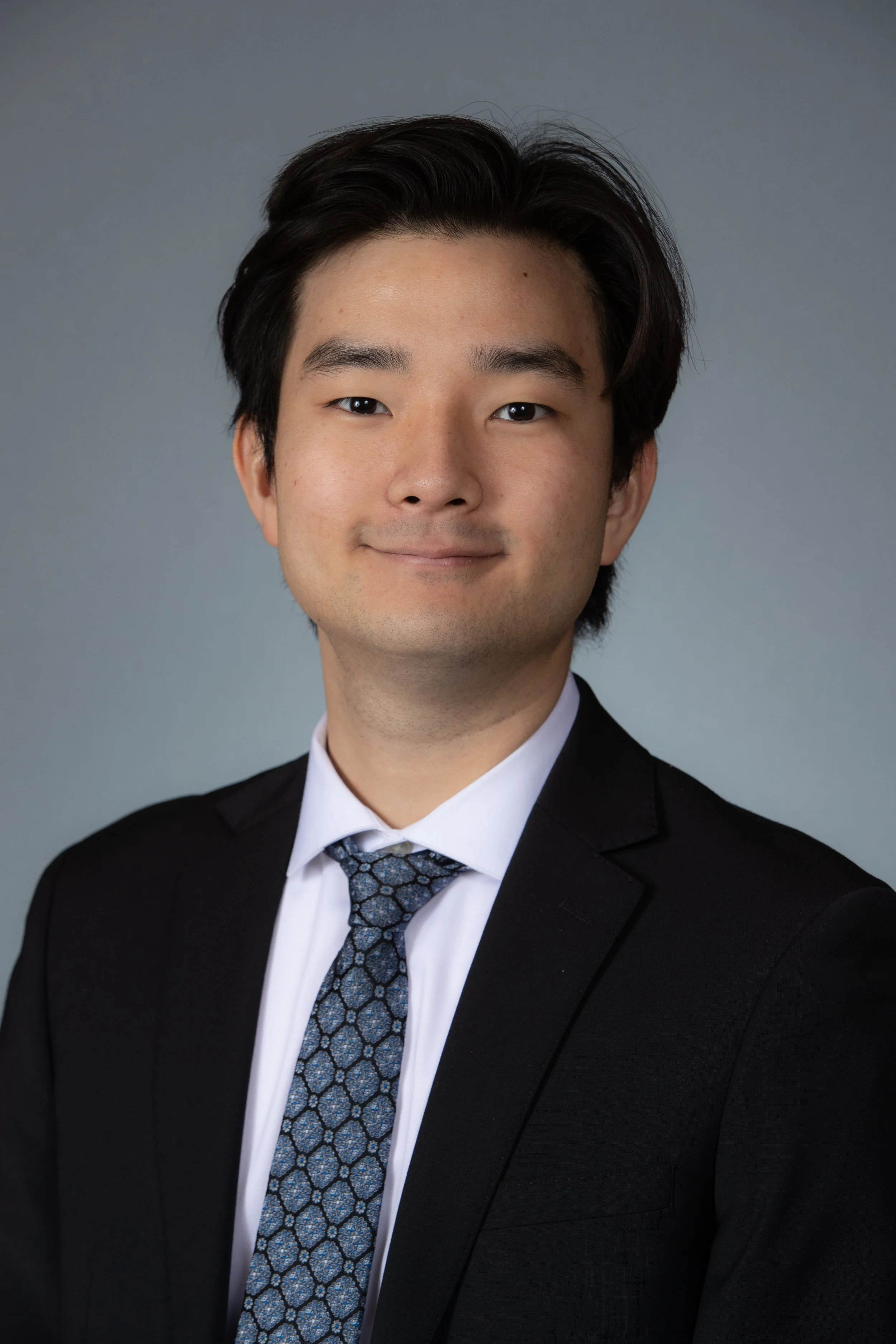 A young man with dark hair wearing a black suit, white shirt, and a patterned tie, smiling softly against a plain gray background.