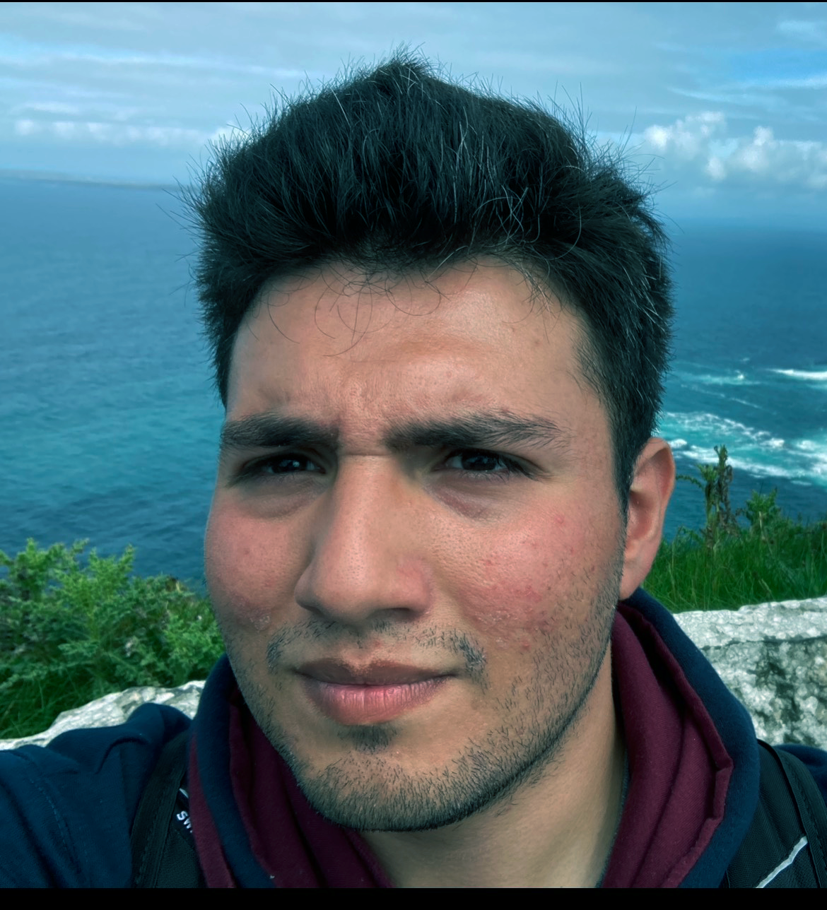 Close-up of a young man with dark hair and facial hair, standing outdoors near the coast with the sea and cloudy sky in the background.
