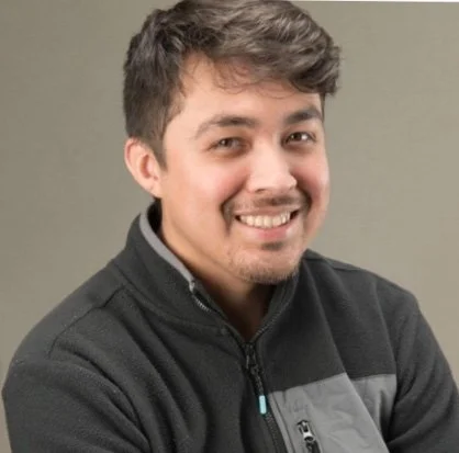 A smiling young man with dark hair, wearing a black jacket with a gray zipper and a gray stripe, standing against a neutral background.