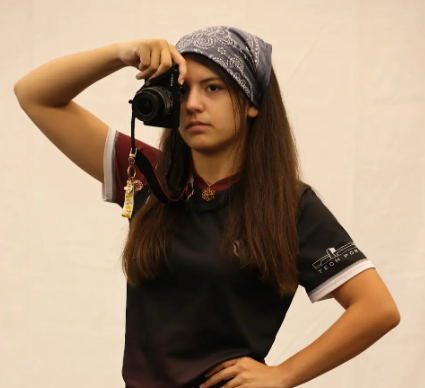 Young woman in a black t-shirt with a gray bandana taking a selfie with a camera in front of a plain background.