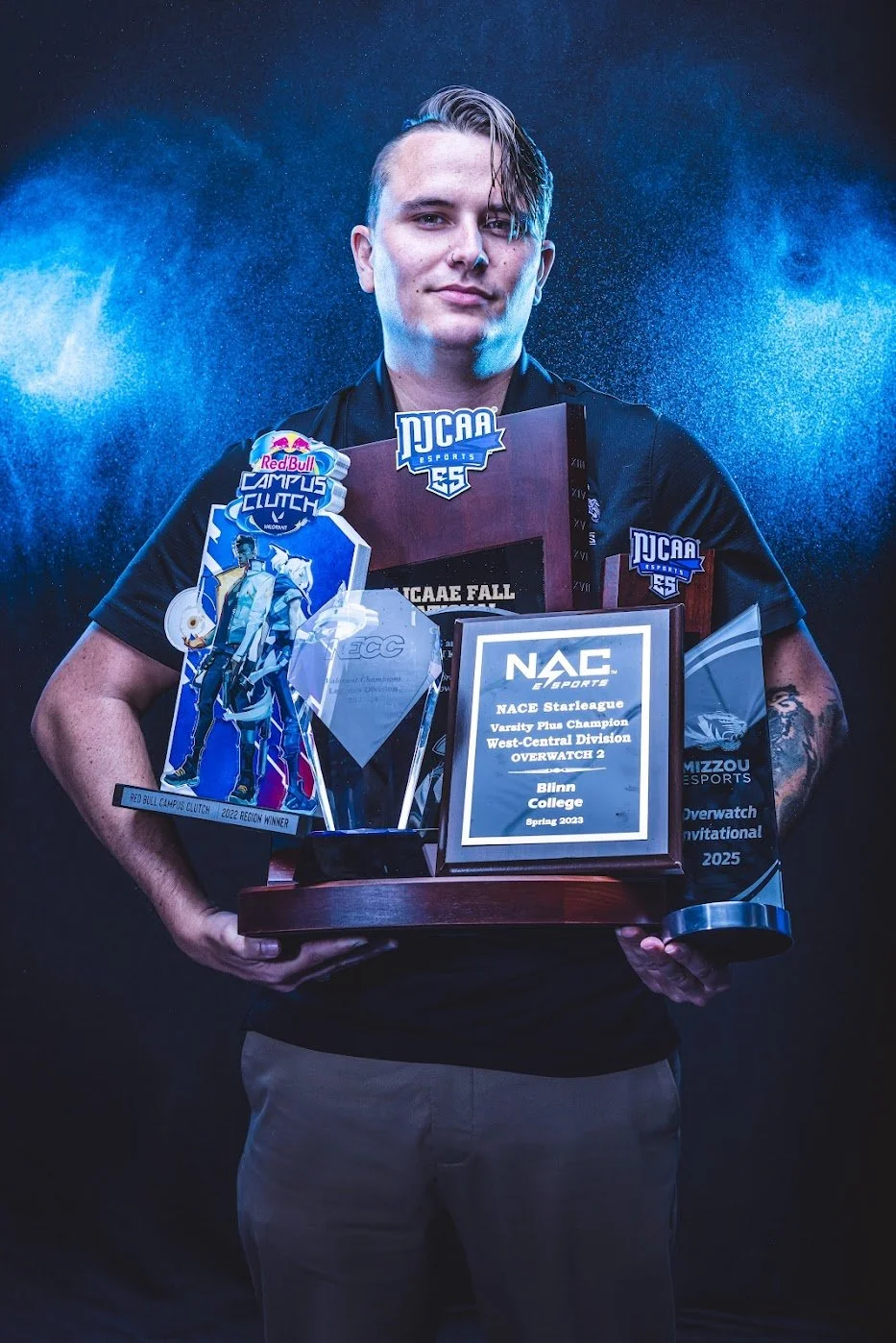 Young man holding multiple awards and trophies from esports competitions, standing against a dark background with blue lighting effects.