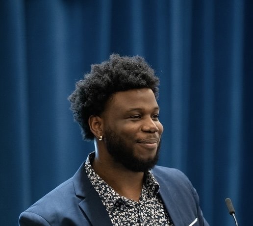 A young Black man with natural hair, wearing a blue blazer and a patterned shirt, smiling in front of a blue curtain.