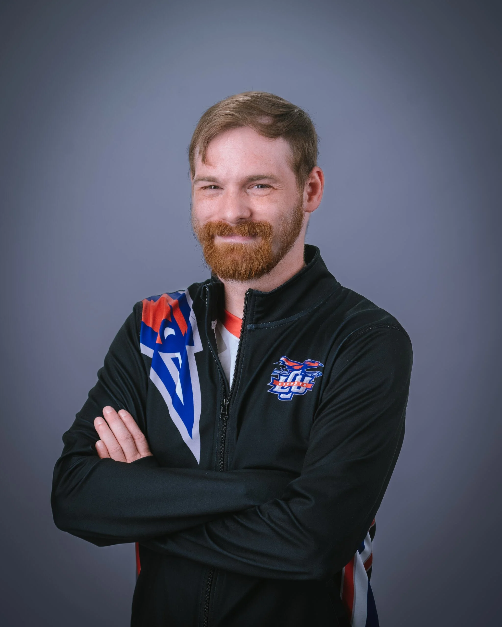 Portrait of a young man with red hair and beard, wearing a black sports jacket with a logo on the chest, standing with arms crossed against a plain gray background, smiling slightly.