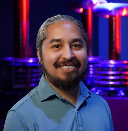 A man with long gray hair and a beard, smiling and wearing a blue collared shirt, standing in front of a colorful, illuminated background.