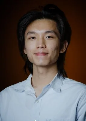 Portrait of a young man with straight black hair, wearing a light blue collared shirt, smiling slightly, against a dark background.