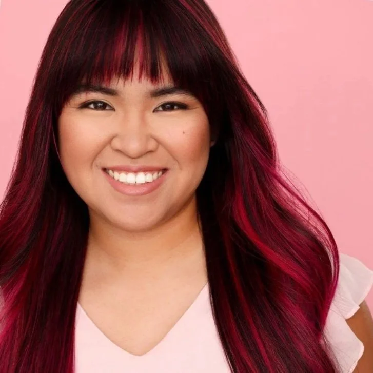 A woman with long, dark red hair and bangs, smiling against a pink background.
