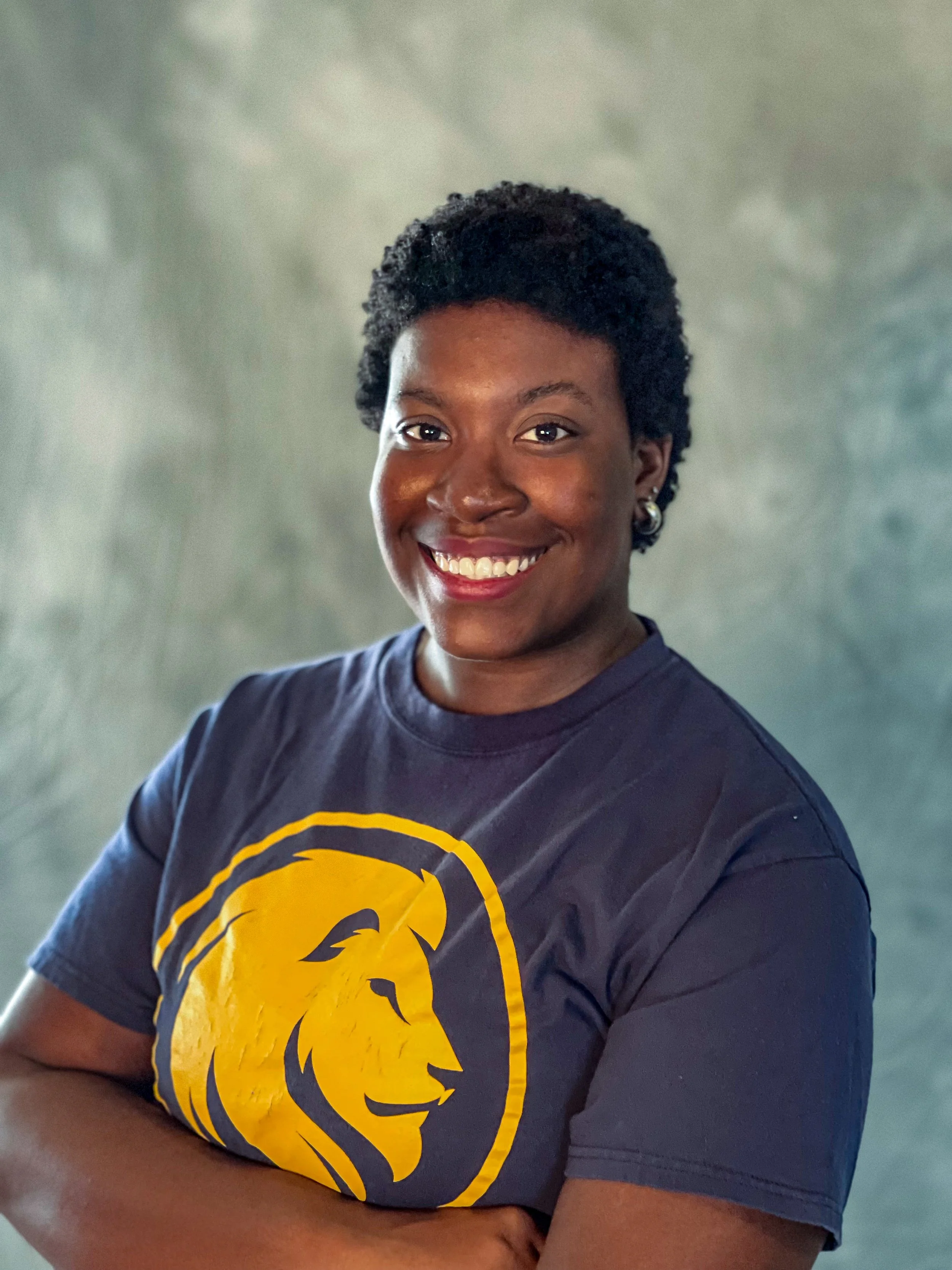 A smiling woman with short curly hair wearing a dark blue T-shirt with a yellow lion logo, standing with arms crossed against a gray background.