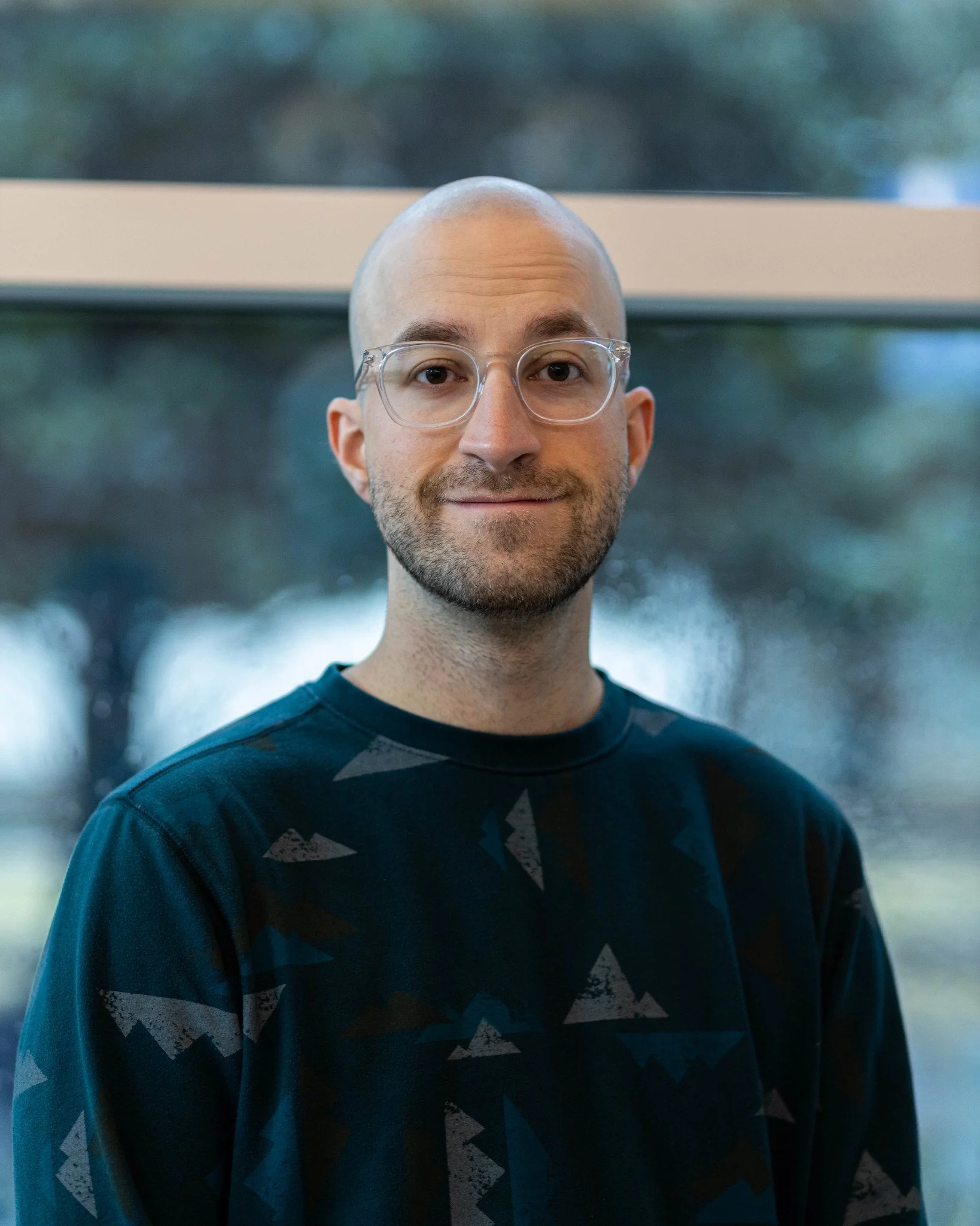 Headshot of a man with glasses, short beard, smiling, standing indoors in front of a large window with trees outside.