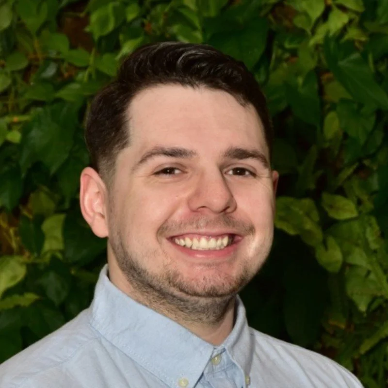 A young man with dark hair and a light beard, smiling broadly, wearing a light-colored button-up shirt, standing in front of green leafy plants.