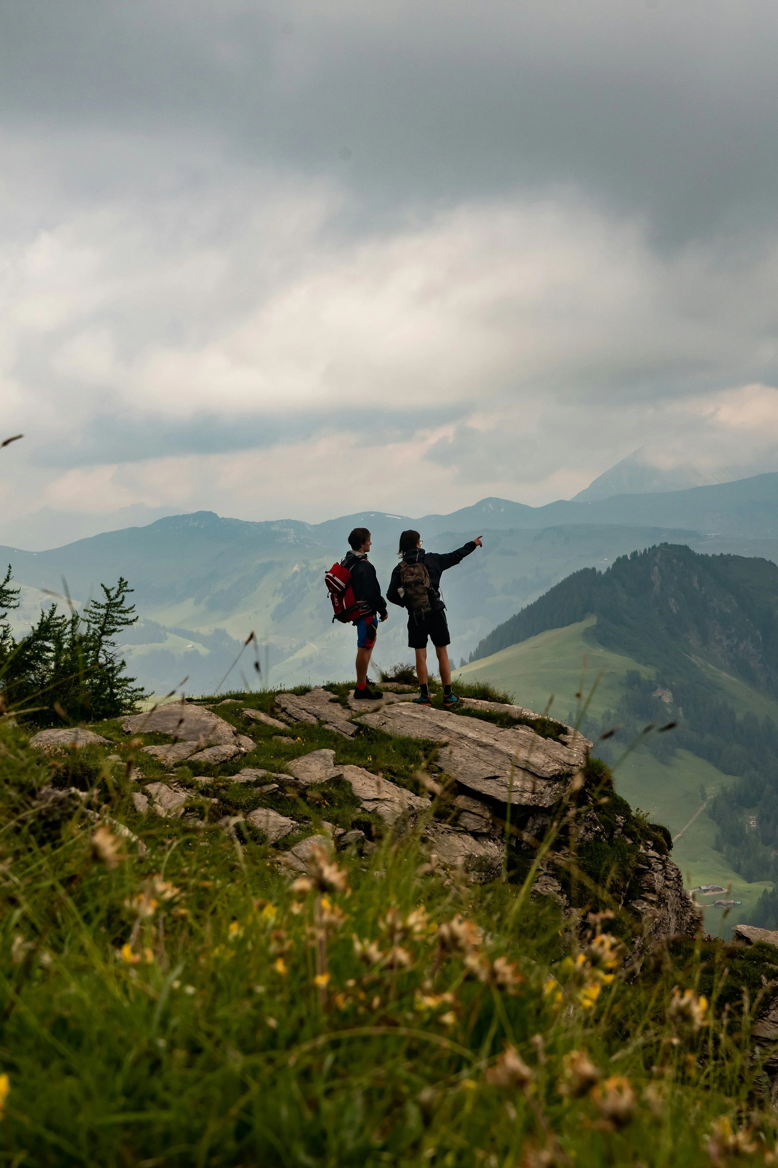 Two hikers standing on rocky terrain, one pointing towards distant mountains in a cloudy sky.