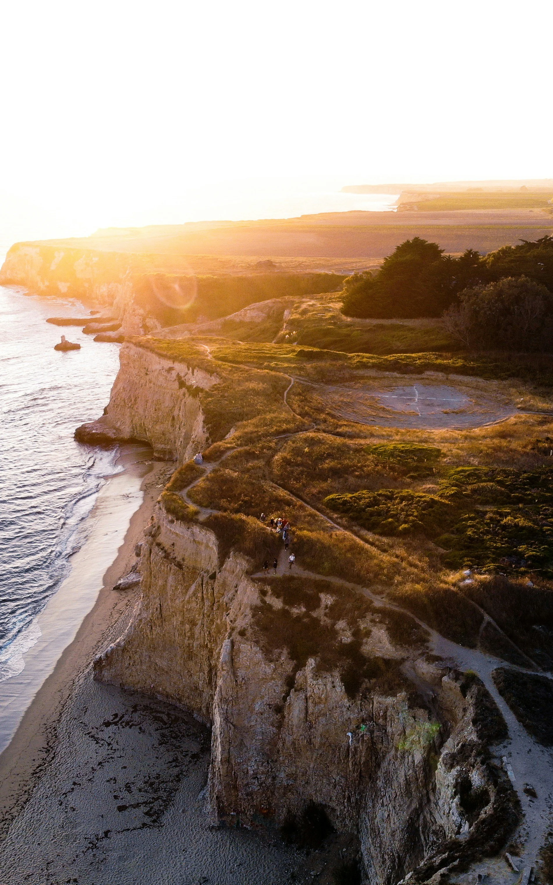 Sunset over rugged coastal cliffs with walking trail and scattered tourists.