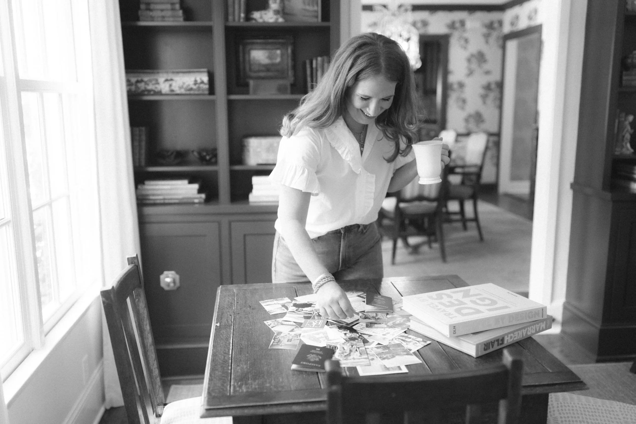 A woman with long hair, smiling, standing at a wooden table, looking at and reaching for a collection of photographs, holding a mug in her other hand, in a room with bookshelves and a dining area in the background.