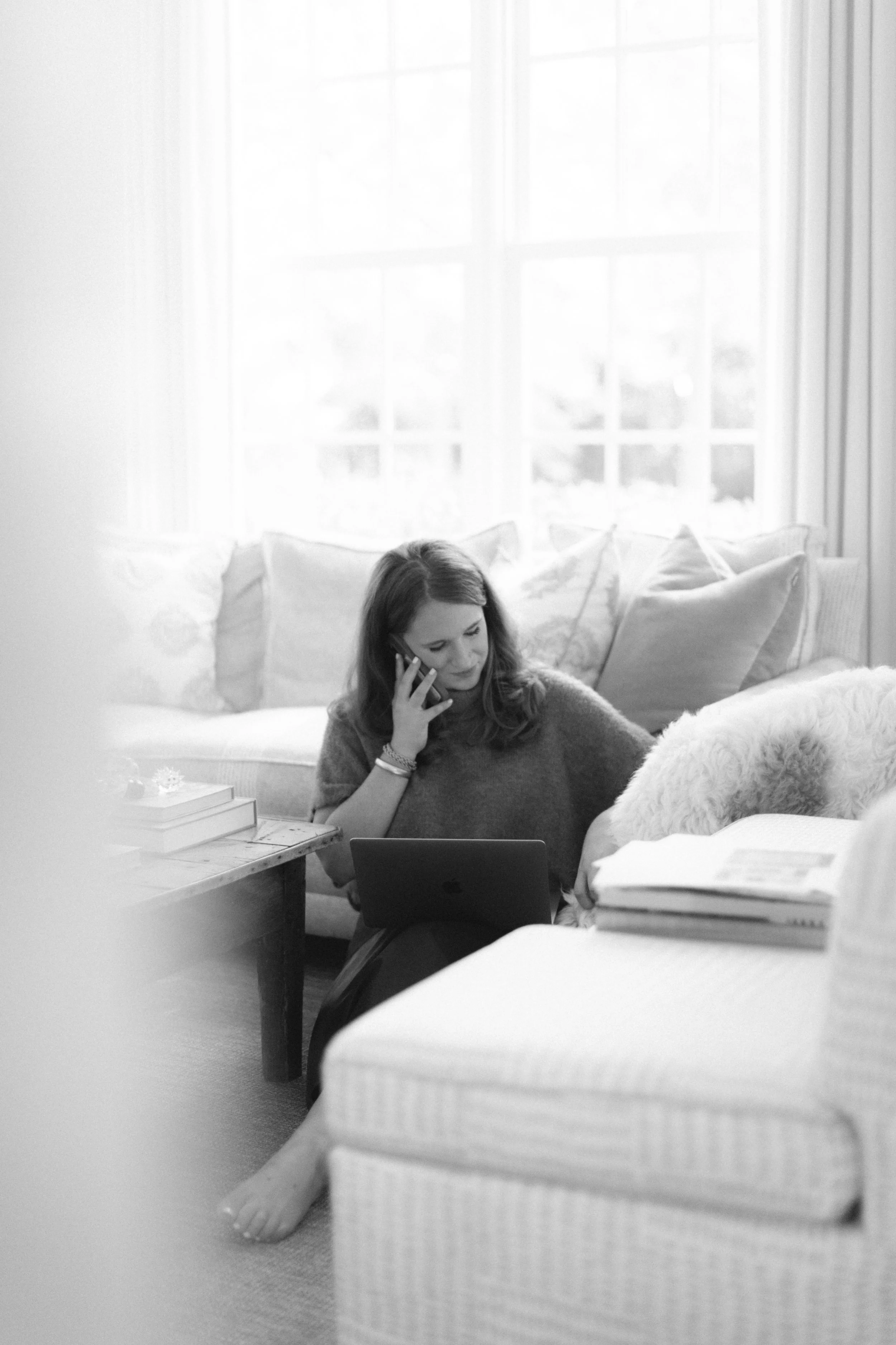 Woman sitting on the floor in a living room, talking on the phone, using a tablet, surrounded by books and pillows.