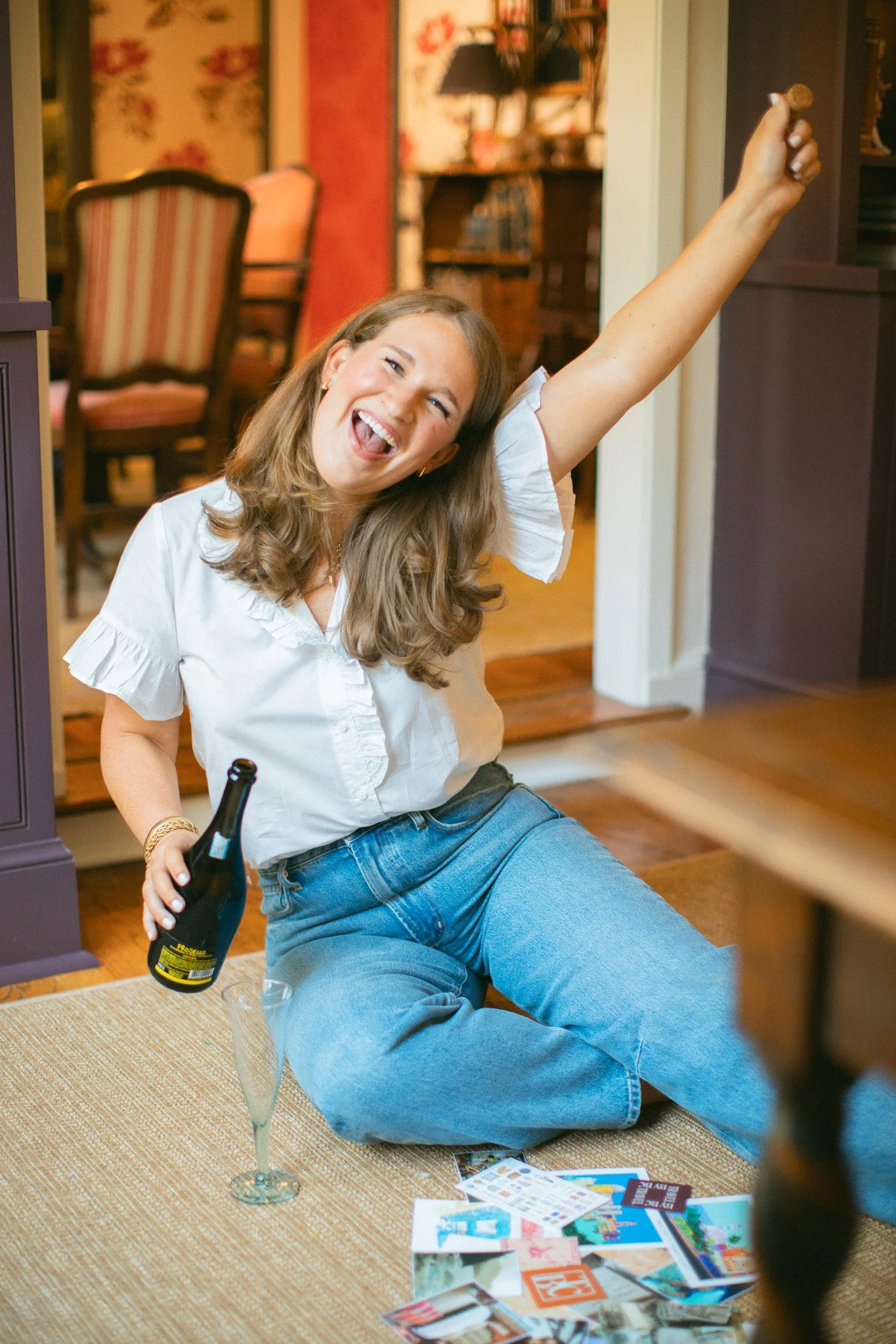A woman sitting on the floor, celebrating, holding a bottle of champagne, with a glass nearby, surrounded by travel brochures and papers, in a cozy home interior.