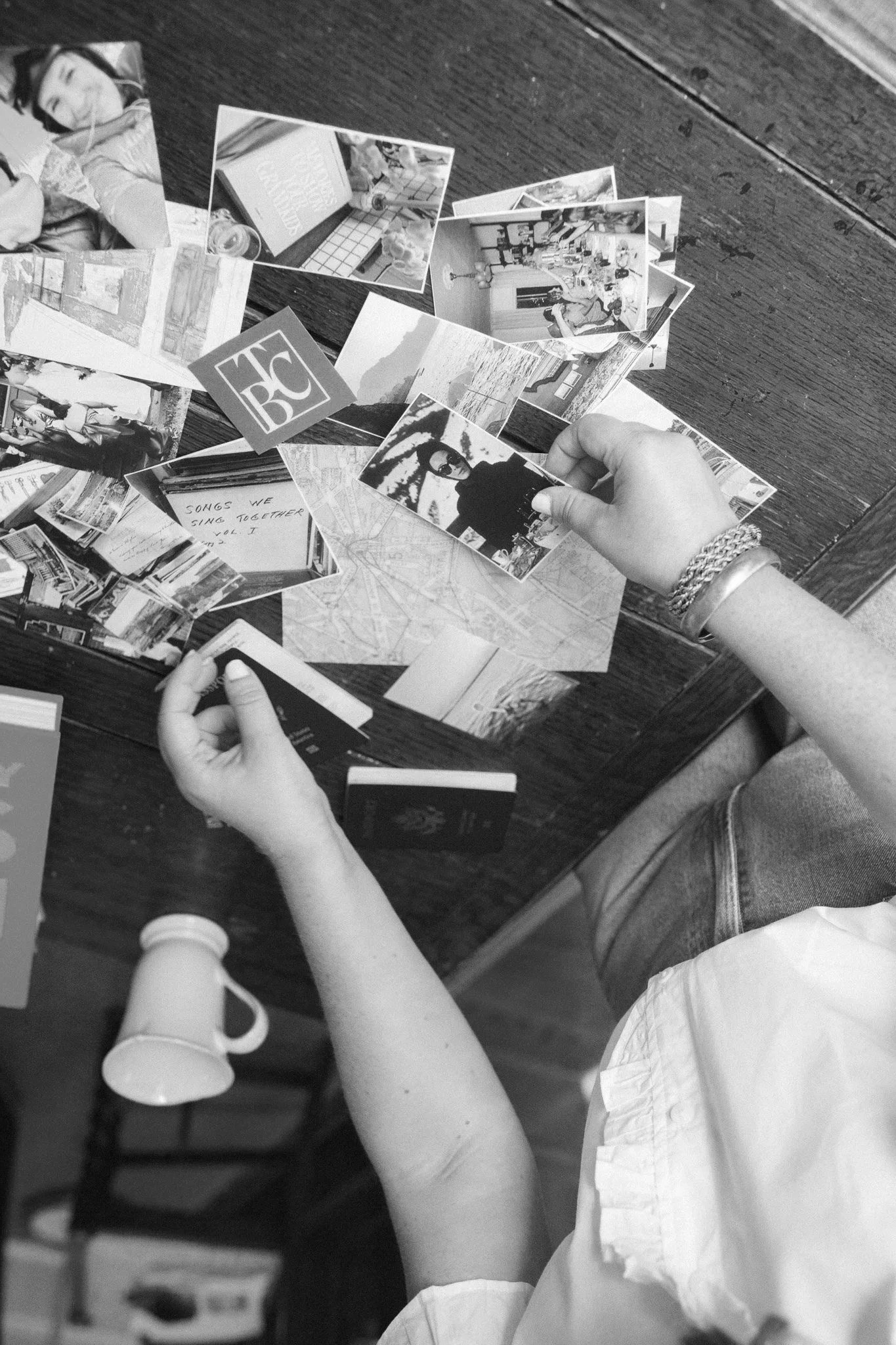 A person with bracelets on their wrist is sitting at a wooden table, looking through a collection of photographs and small printed images spread out before them.