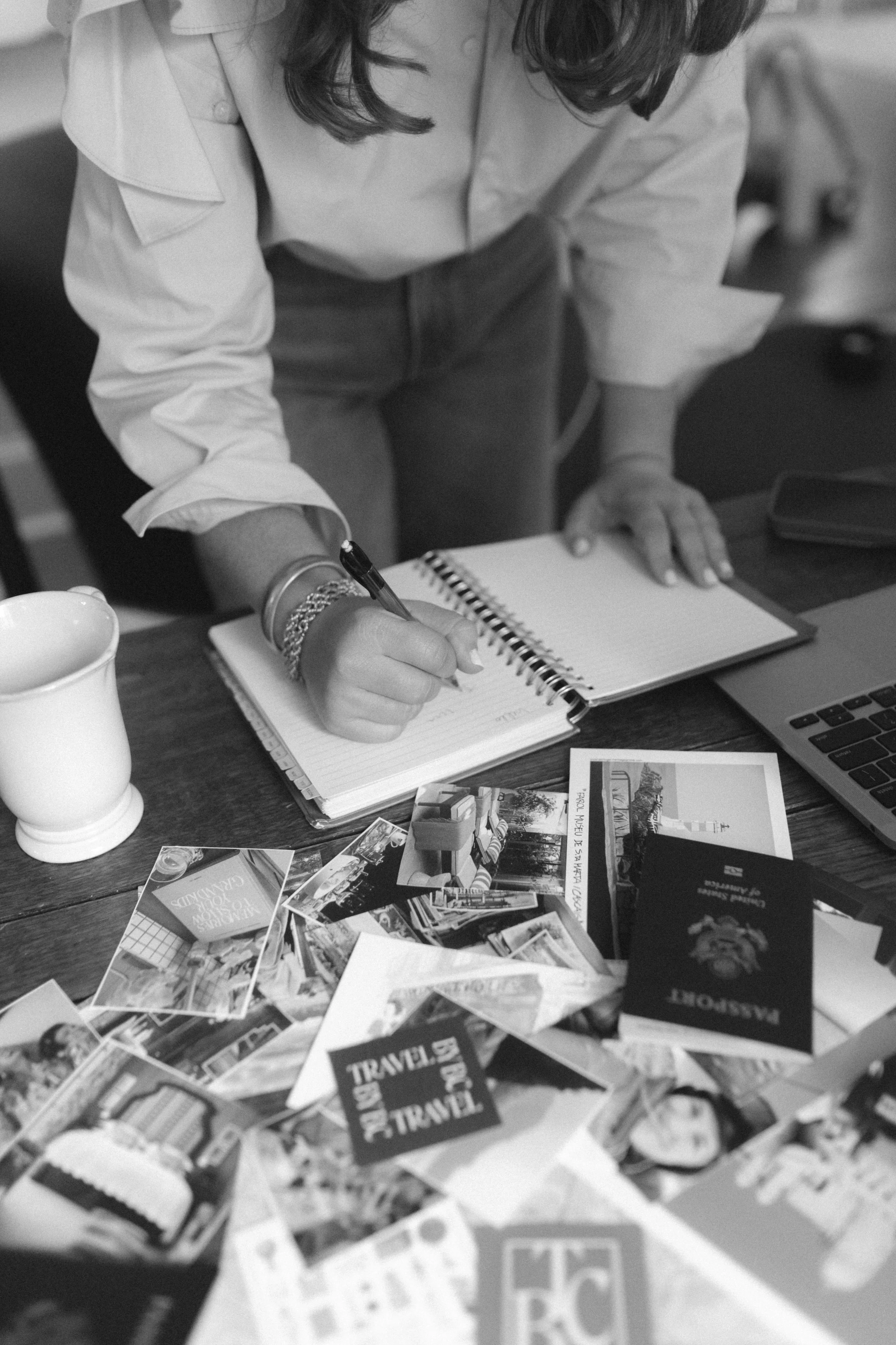 A person writing in a notebook on a table cluttered with travel photos, postcards, and a passport, with a mug, laptop, and other items nearby.